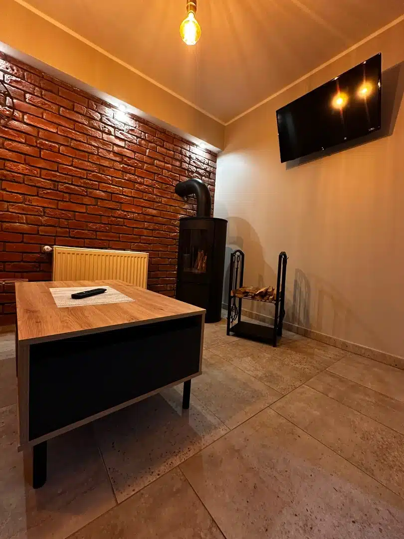 Salon w apartamencie Legia, Vigor Horses, z ciepłym oświetleniem, piecem kominkowym, ceglaną ścianą i telewizorem, zachęcający do relaksu w przytulnej atmosferze