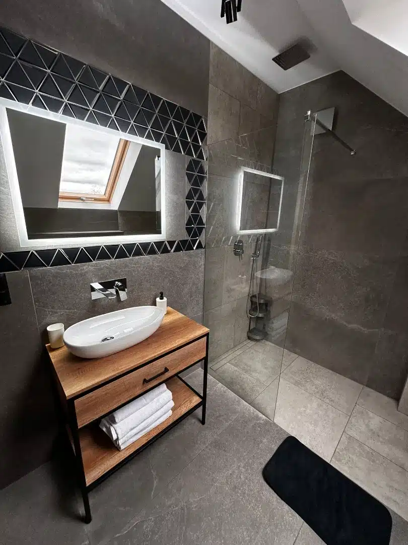 Widok na przestronną łazienkę w apartamencie Karino, Vigor Horses, ukazujący prysznic bez kabiny, umywalkę na drewnianej szafce i okno dachowe zapewniające naturalne oświetlenie