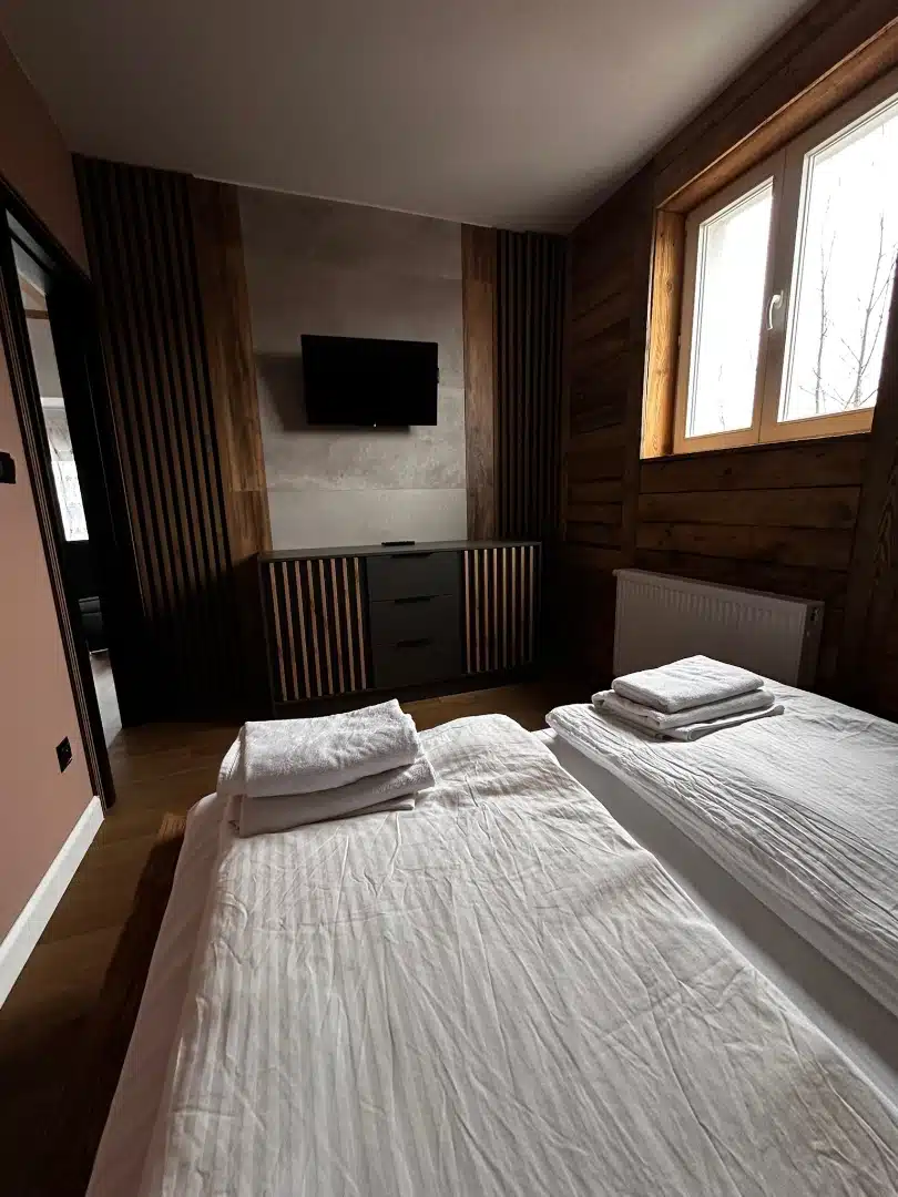 Przytulna sypialnia z dwoma pojedynczymi łóżkami w apartamencie Karino, Vigor Horses, ozdobiona drewnianymi elementami i nowoczesnym telewizorem na ścianie