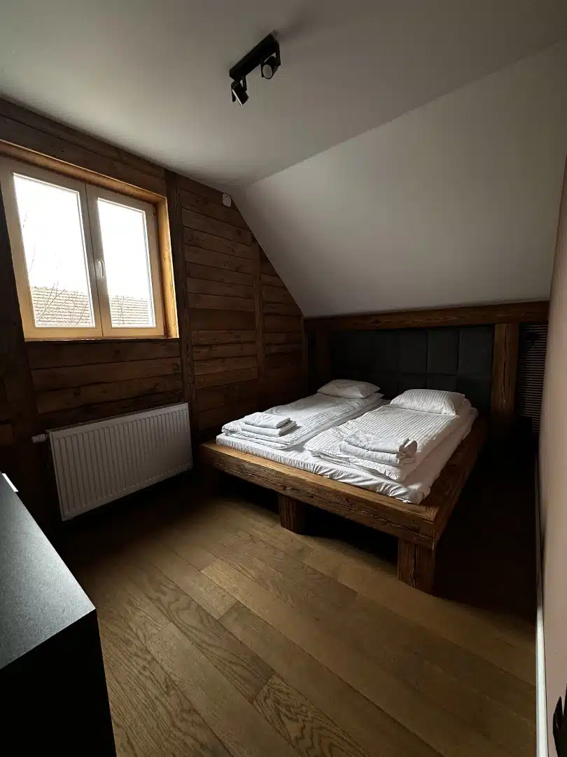 Widok na podwójne łóżko w sypialni apartamentu Karino, Vigor Horses, uchwycony z innej perspektywy, podkreślający drewniane wykończenia i przytulny charakter pomieszczenia
