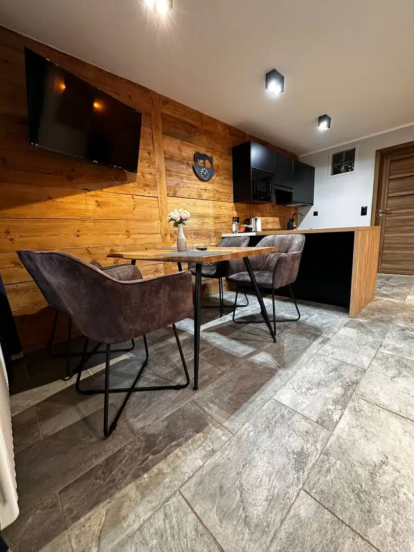 Funkcjonalna przestrzeń jadalniana z kuchnią w apartamencie Legia, Vigor Horses, wyposażona w drewniany stół z eleganckimi krzesłami i telewizorem na ścianie, tworząca przytulną atmosferę do wspólnych posiłków