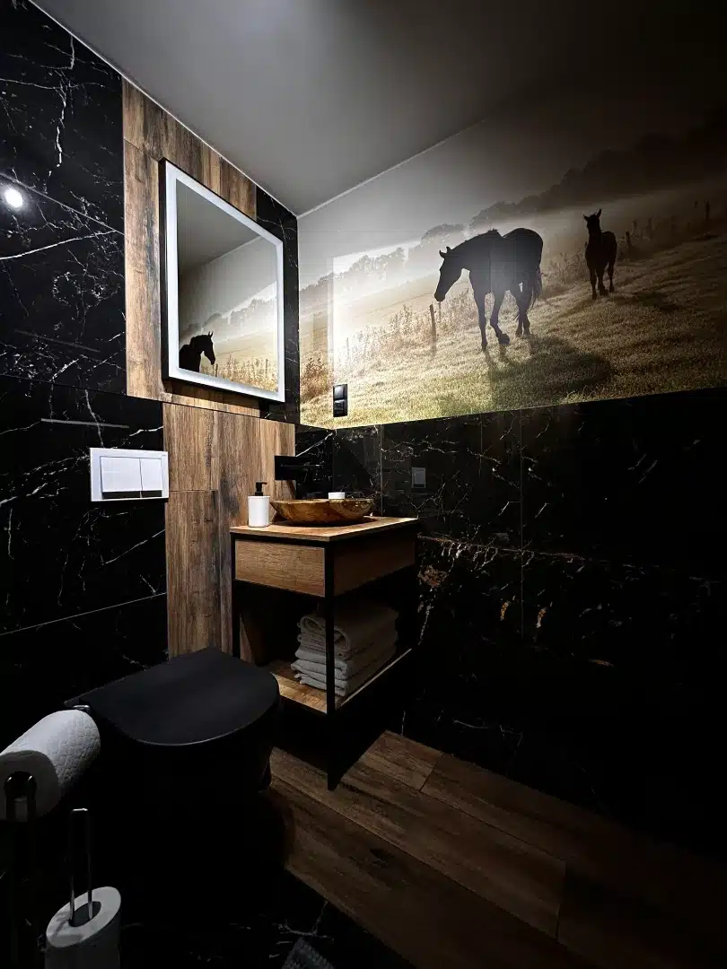 Zakątek z umywalką w łazience apartamentu Legia, Vigor Horses, z efektownym obrazem pastwisk z końmi w tle, marmurowymi ścianami i drewnianym blatem umywalki, tworzącym harmonijne połączenie natury z nowoczesnością