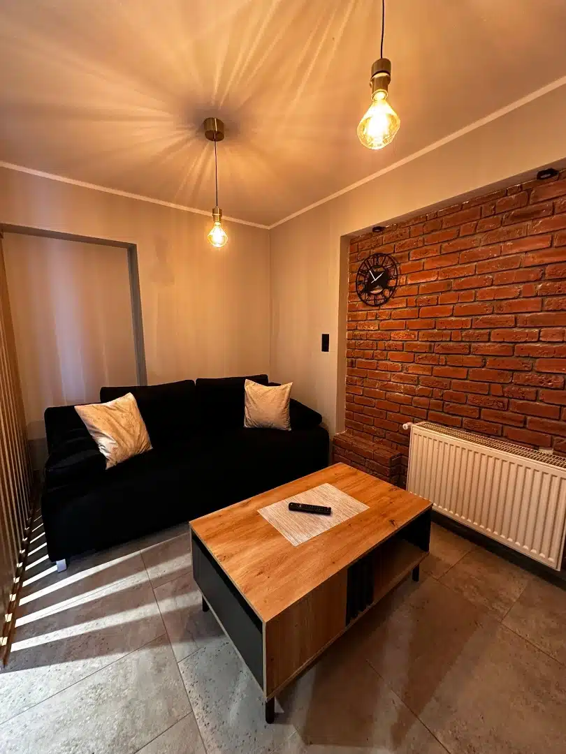 Salon w apartamencie Legia, Vigor Horses, z przytulnym czarnym narożnikiem, drewnianym stolikiem kawowym i ceglany ścianą z zegarem, tworzący ciepłą i gościnną atmosferę
