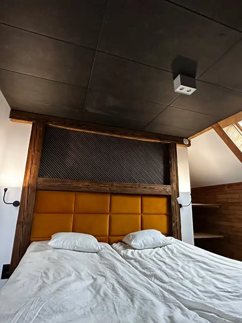 Przytulna sypialnia w apartamencie Merida, Vigor Horses, z dużym łóżkiem z żółtym wezgłowiem, drewnianymi ścianami i oknem dachowym wpuszczającym naturalne światło