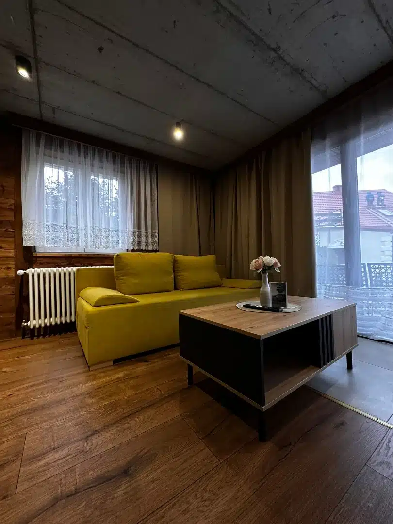 Salon apartamentu Ursa, Vigor Horses, ożywiony jaskrawo żółtą sofą, drewnianą podłogą i delikatnymi, białymi firankami, tworzący przytulną atmosferę do relaksu i spotkań