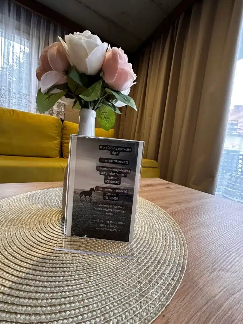 Stół w apartamencie Ursa, Vigor Horses, z eleganckim stojakiem informacyjnym i bukietem sztucznych róż, zapraszającym gości do spędzenia czasu w komfortowych warunkach